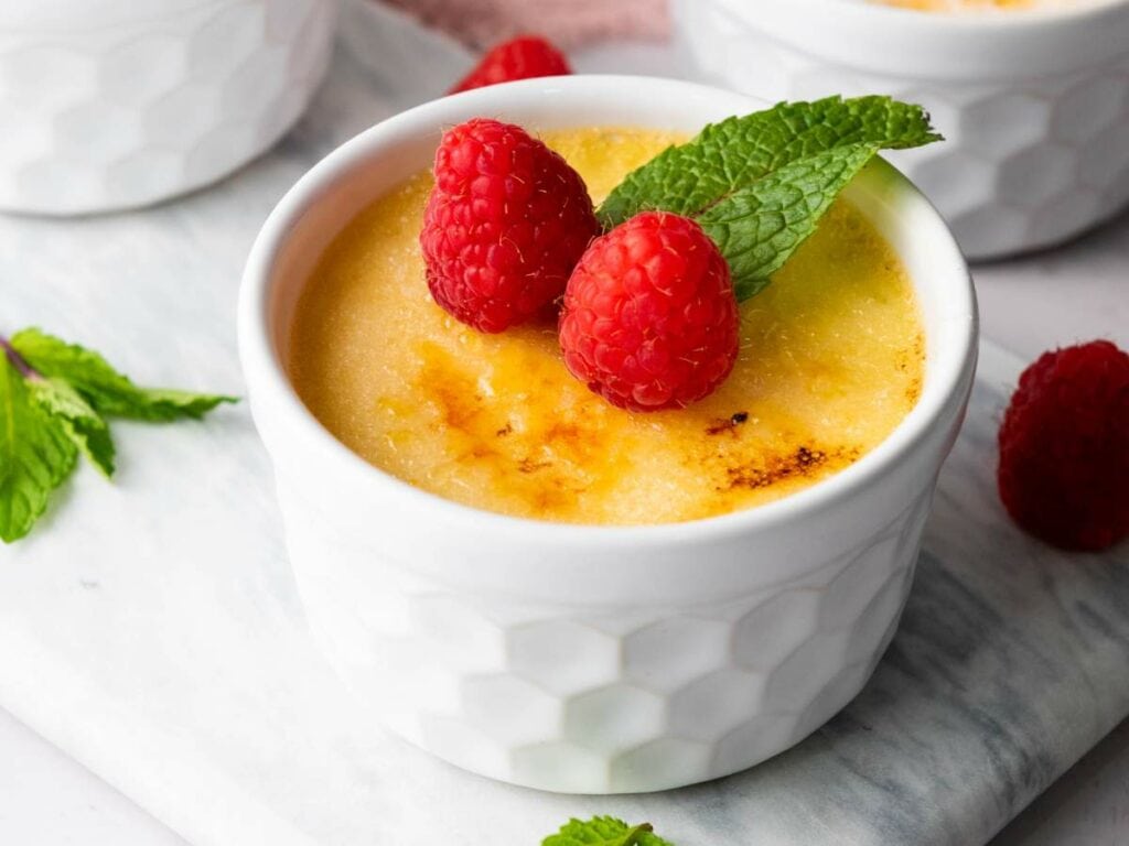 A ramekin of crème brûlée topped with two raspberries and a mint leaf, placed on a white surface.