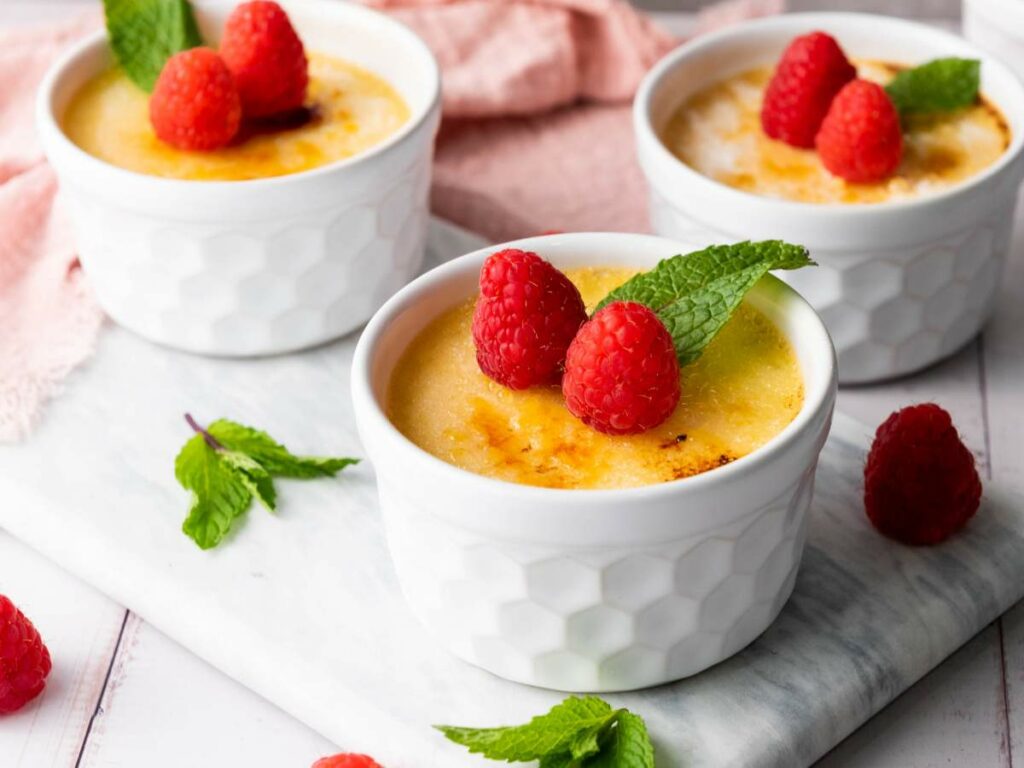 Three white ramekins filled with burnt creme, each topped with two raspberries and a mint leaf.
