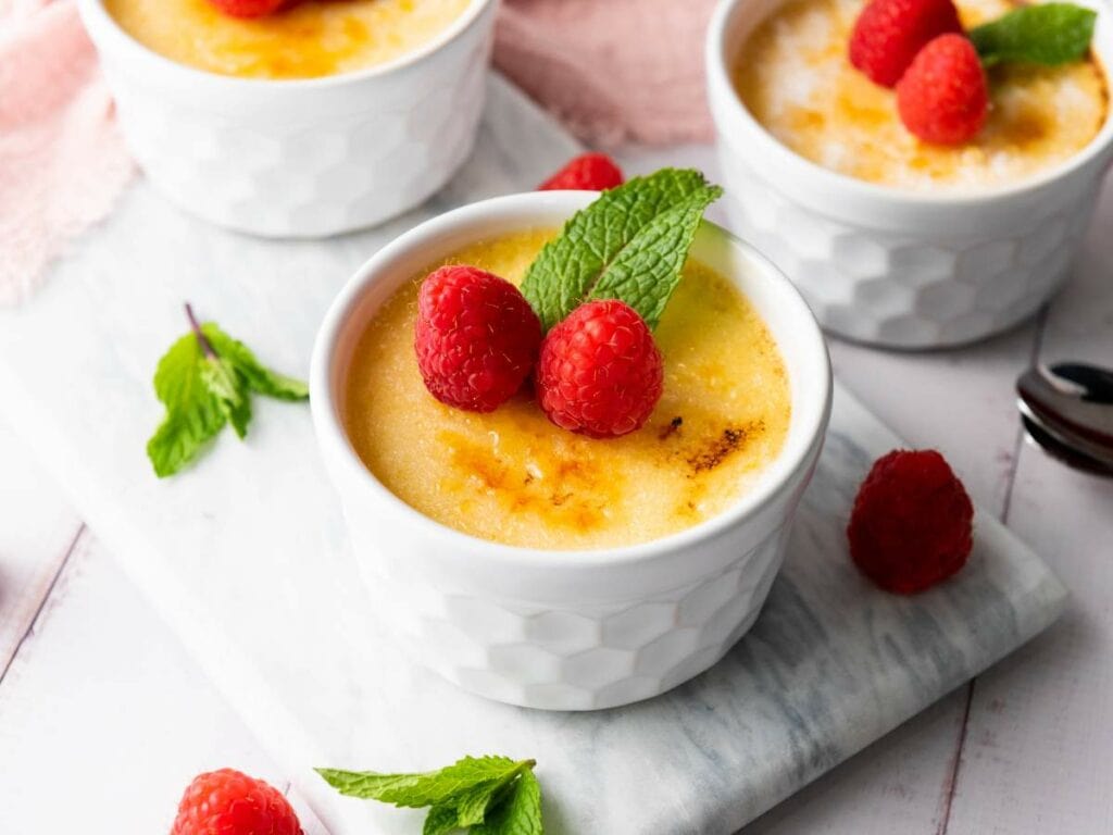 A white ramekin filled with crème brûlée, topped with fresh raspberries and a mint leaf, sits on a marble surface.