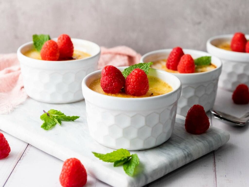 Four white ramekins filled with custard, each topped with fresh raspberries and mint leaves.