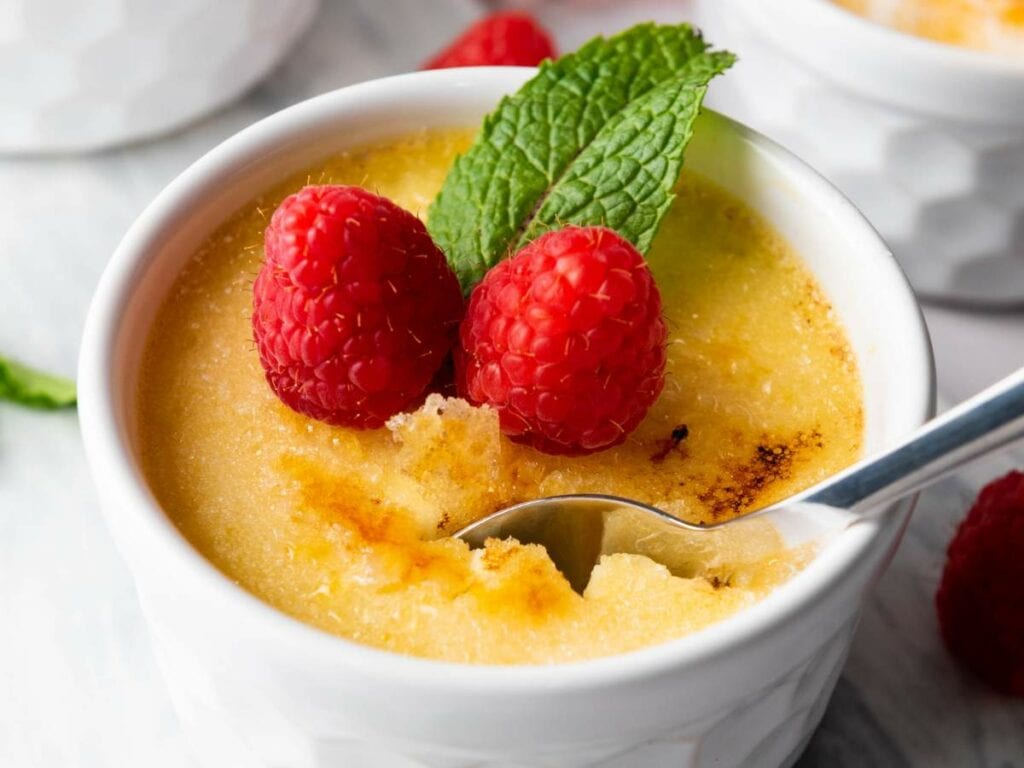 A white ramekin filled with crème brûlée, topped with two raspberries and a mint leaf, with a spoon breaking the caramelized sugar crust.