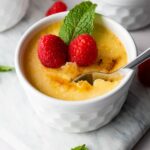 A white ramekin filled with crème brûlée, topped with two raspberries and a mint sprig, with a spoon scooping out a portion.