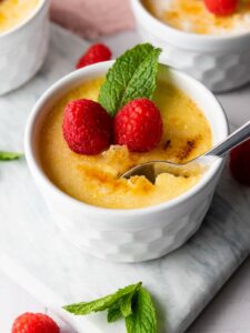 A white ramekin filled with crème brûlée, topped with two raspberries and a mint sprig, with a spoon scooping out a portion.