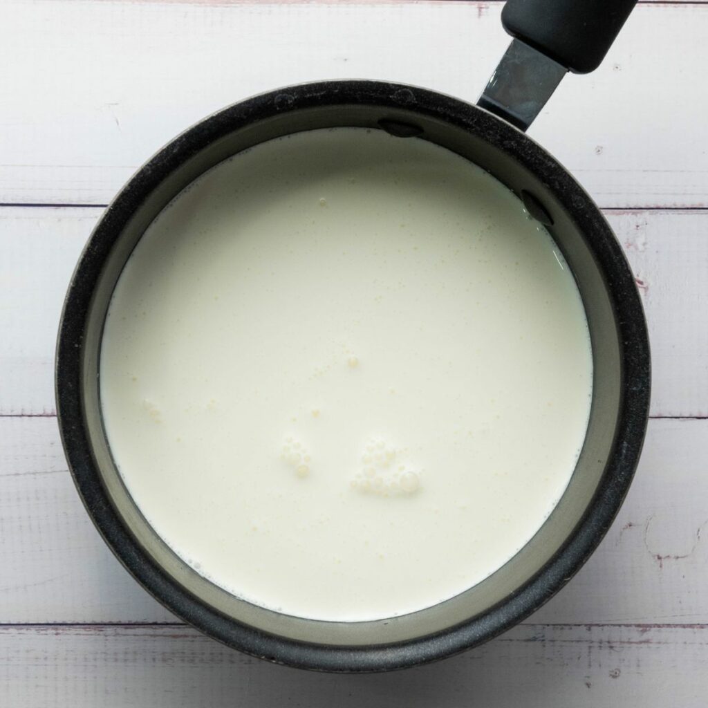 A black saucepan filled with heavy cream sits on a white wooden surface.