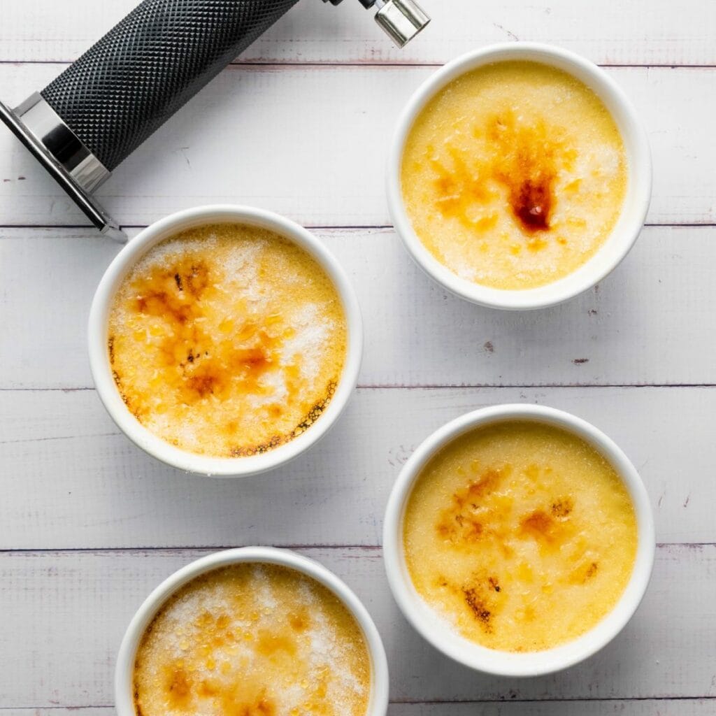 Four ramekins of crème brûlée with caramelized tops sit on a white wooden surface next to a kitchen torch.