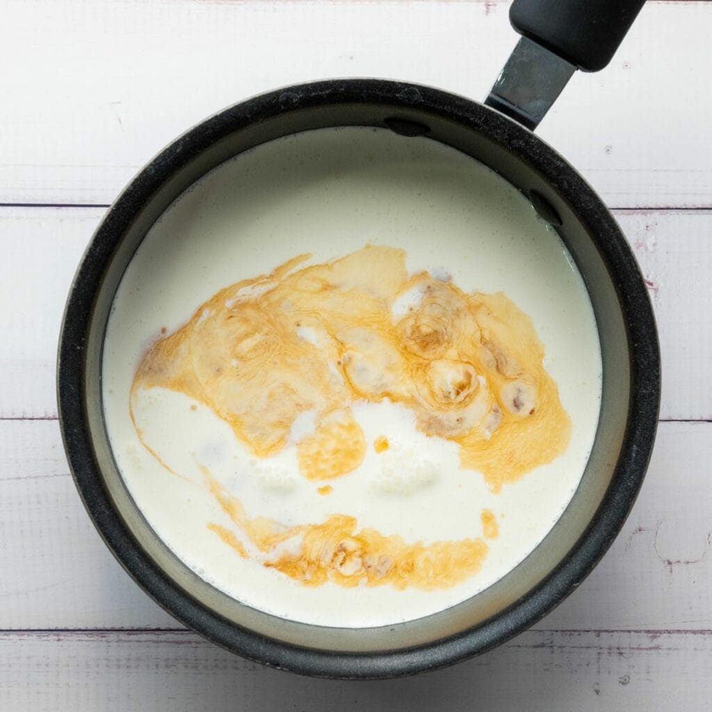 A saucepan with cream and vanilla extract swirling together on a white wooden surface.