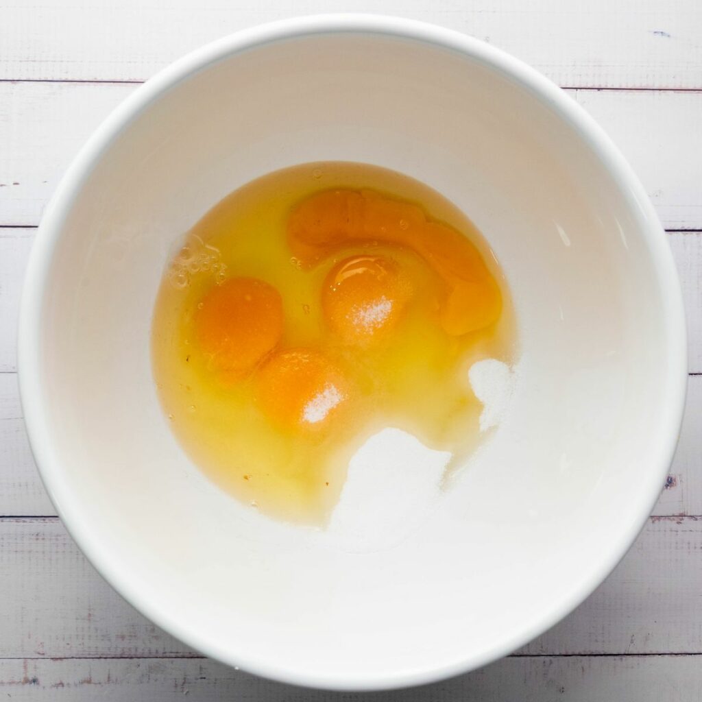 A white bowl containing cracked raw eggs with visible yolks, sugar and salt on a white wooden surface.