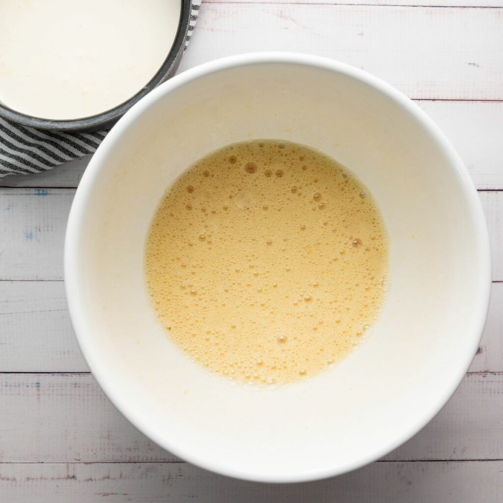 A large white bowl containing a frothy, light yellow liquid mixture sits on a white wooden surface.