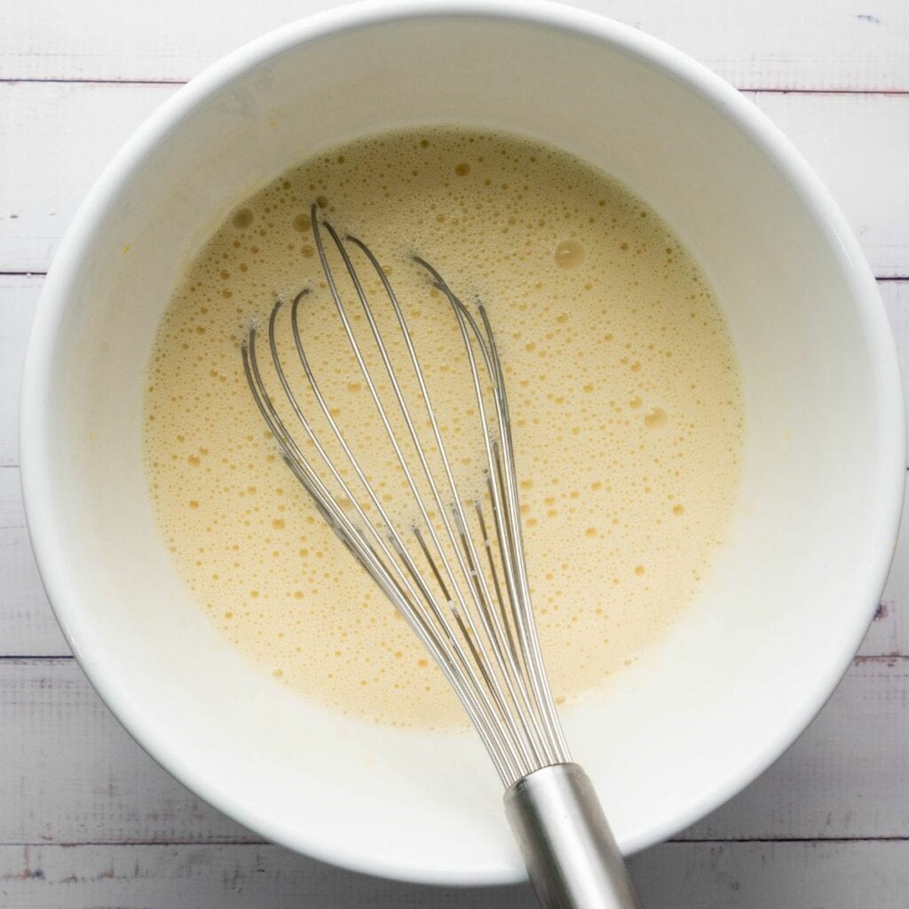 A metal whisk rests in a white bowl filled with a light yellow, frothy liquid mixture on a white surface.
