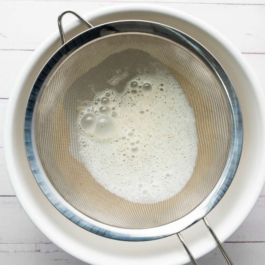 A metal strainer with foam and liquid sits over a white bowl on a light-colored surface.