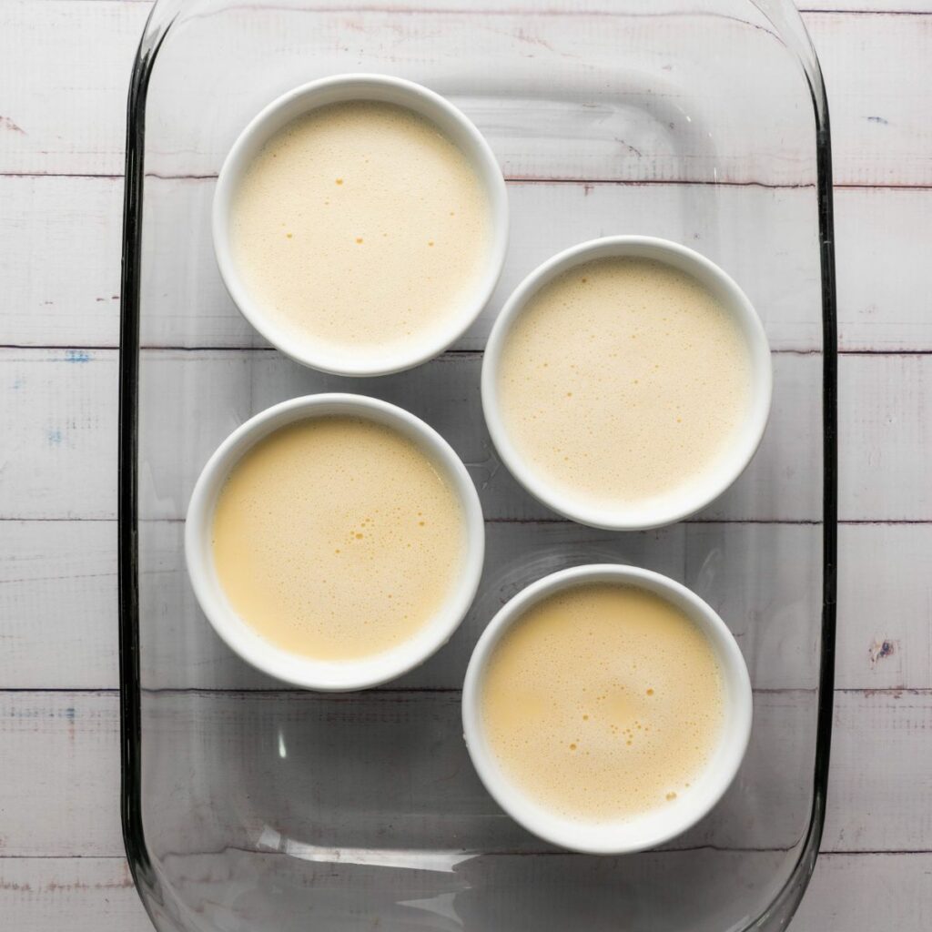 Four white ramekins filled with a light yellow mixture are placed in a glass baking dish on a white wooden surface.