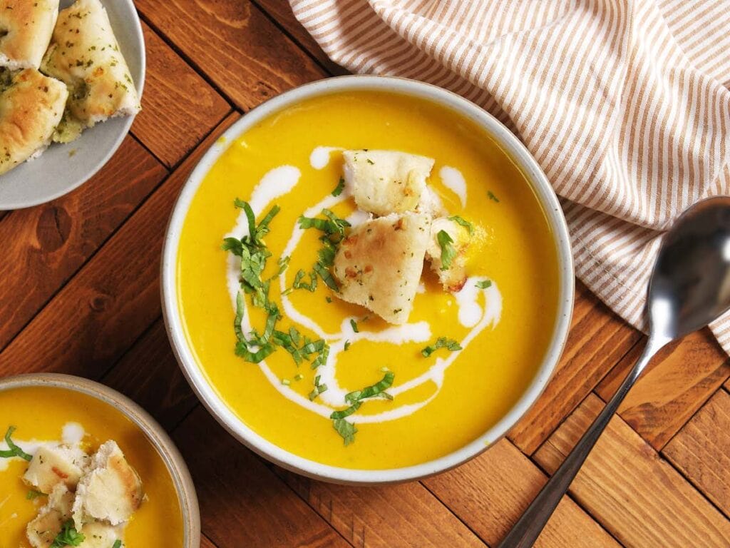 A bowl of creamy yellow soup garnished with swirls of cream, chopped herbs, and pieces of flatbread, on a wooden table next to a striped cloth and a spoon.