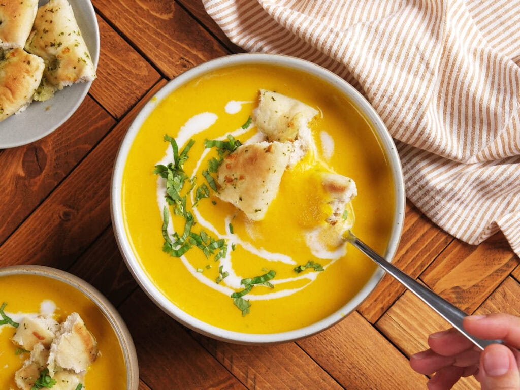 A bowl of creamy yellow soup garnished with herbs and cream, topped with pieces of bread, with a hand holding a spoon.