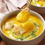 A bowl of creamy yellow soup garnished with chopped herbs, flatbread pieces, and a swirl of cream, with a spoonful of soup being lifted above the bowl.