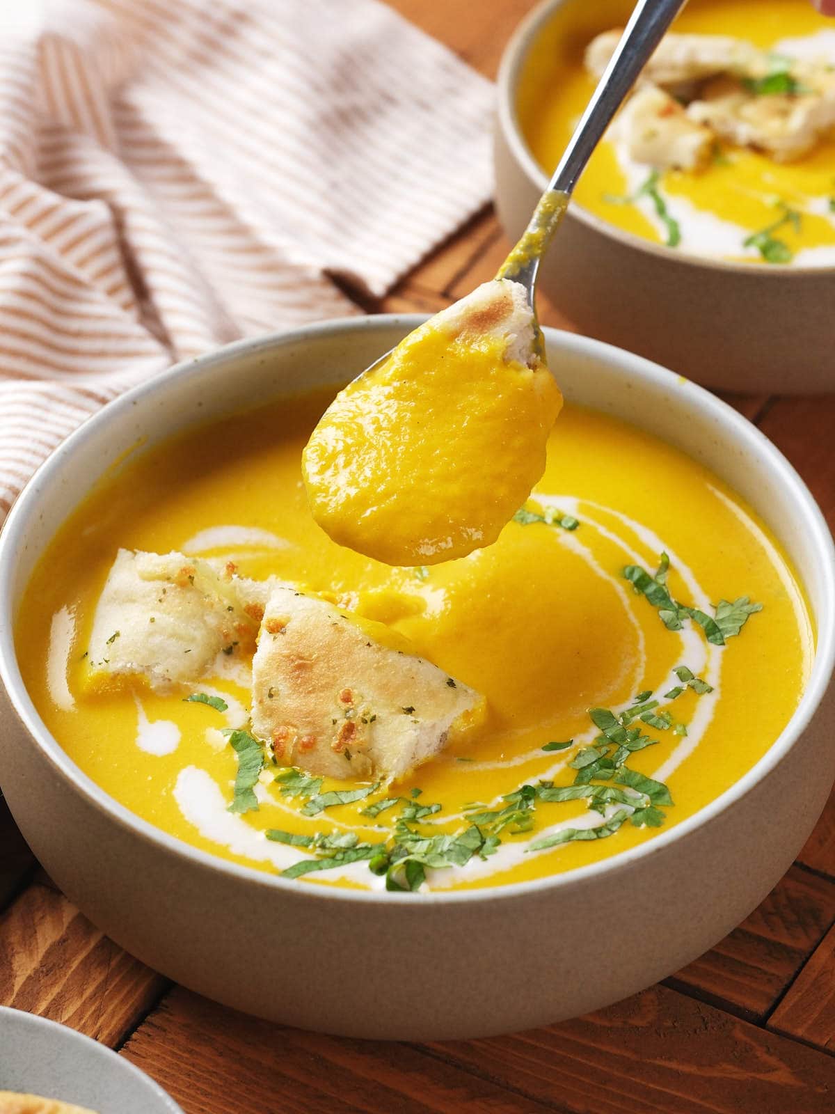 A bowl of creamy yellow soup garnished with chopped herbs, flatbread pieces, and a swirl of cream, with a spoonful of soup being lifted above the bowl.