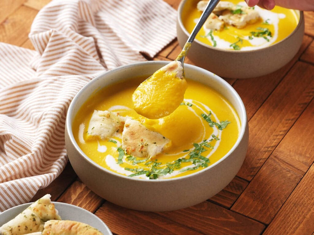 A bowl of creamy yellow soup garnished with herbs and pieces of bread, with a spoon lifting a portion; another bowl and a striped cloth are nearby.