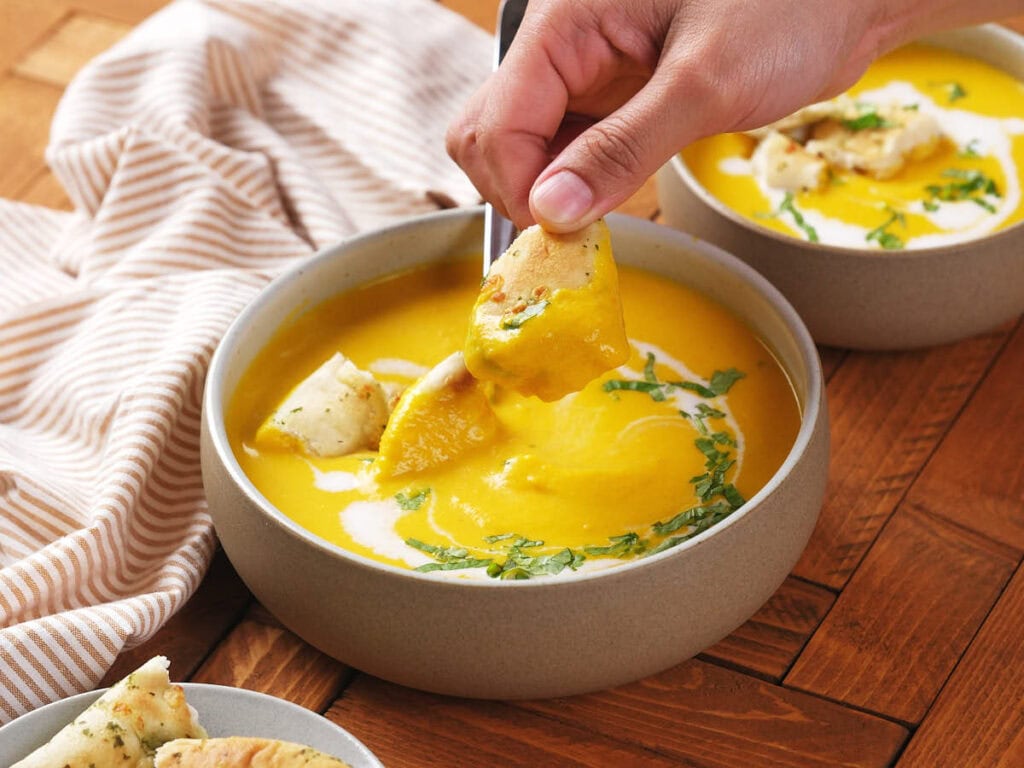 A hand dips a piece of bread into a bowl of creamy orange soup garnished with herbs.