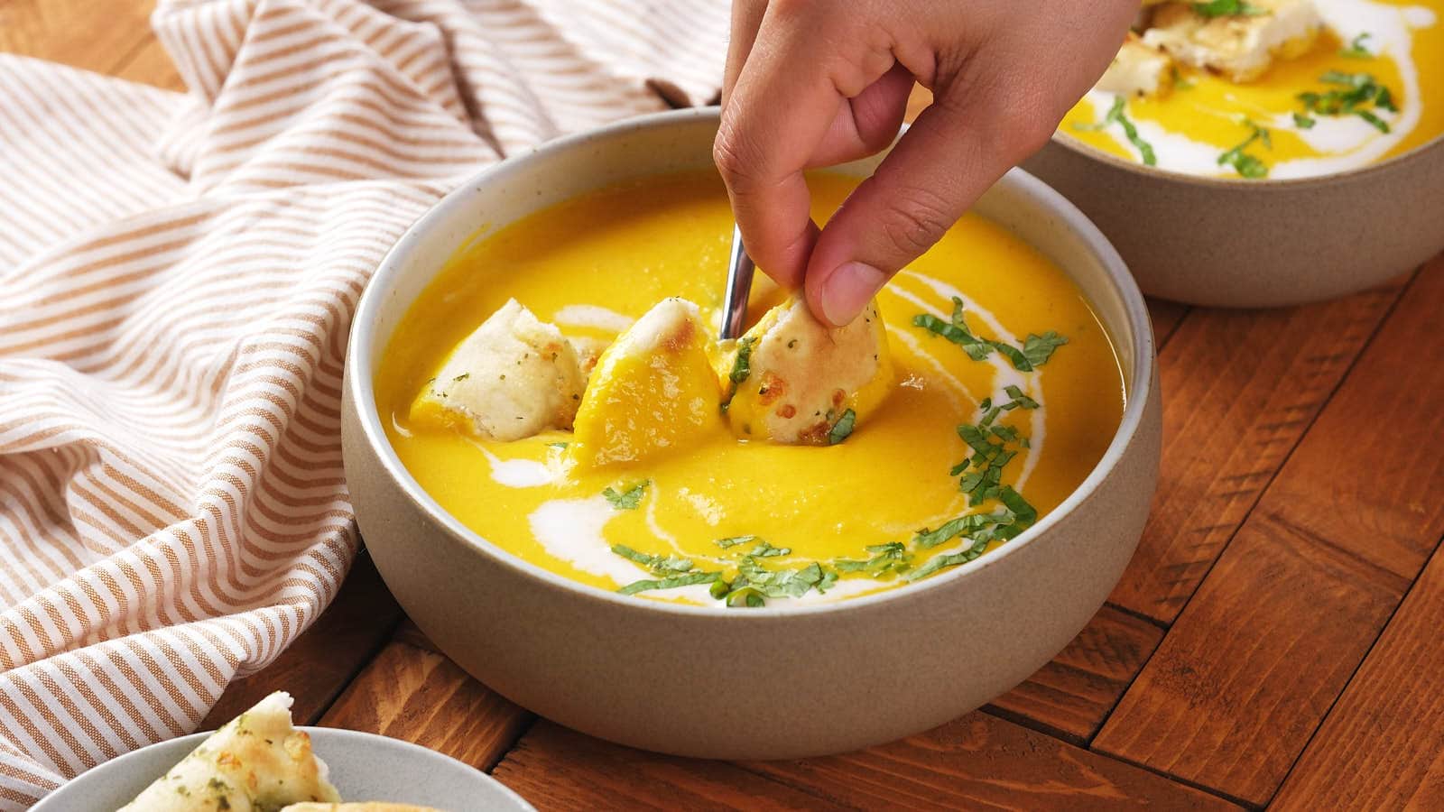 A hand dips bread into a bowl of creamy yellow soup garnished with herbs and a swirl of cream, on a wooden table.