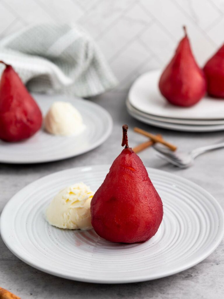 A red pear with a scoop of vanilla ice cream on a white plate, with more poached pears and plates in the background.