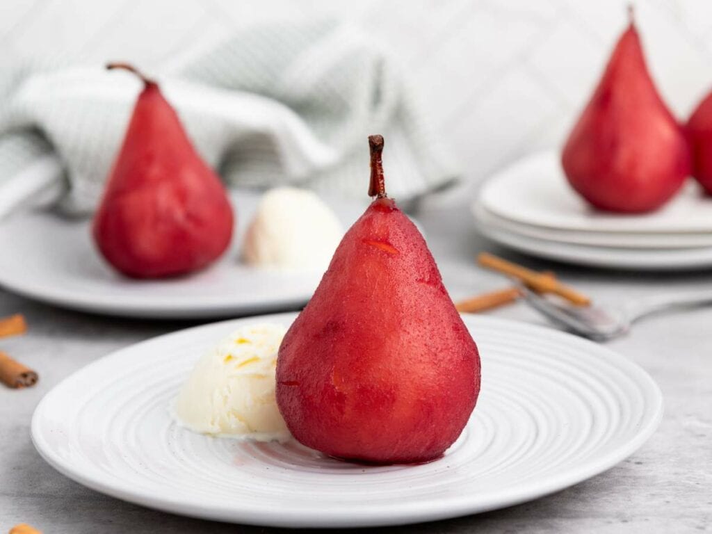 A poached pear with a scoop of vanilla ice cream is served on a white plate, with another plate of the same dessert in the background.