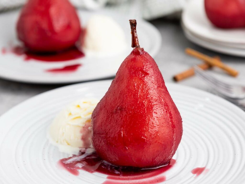 A poached pear in red sauce is served on a white plate with a scoop of vanilla ice cream.