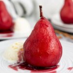 A poached pear in red wine sauce served on a white plate with a scoop of vanilla ice cream.