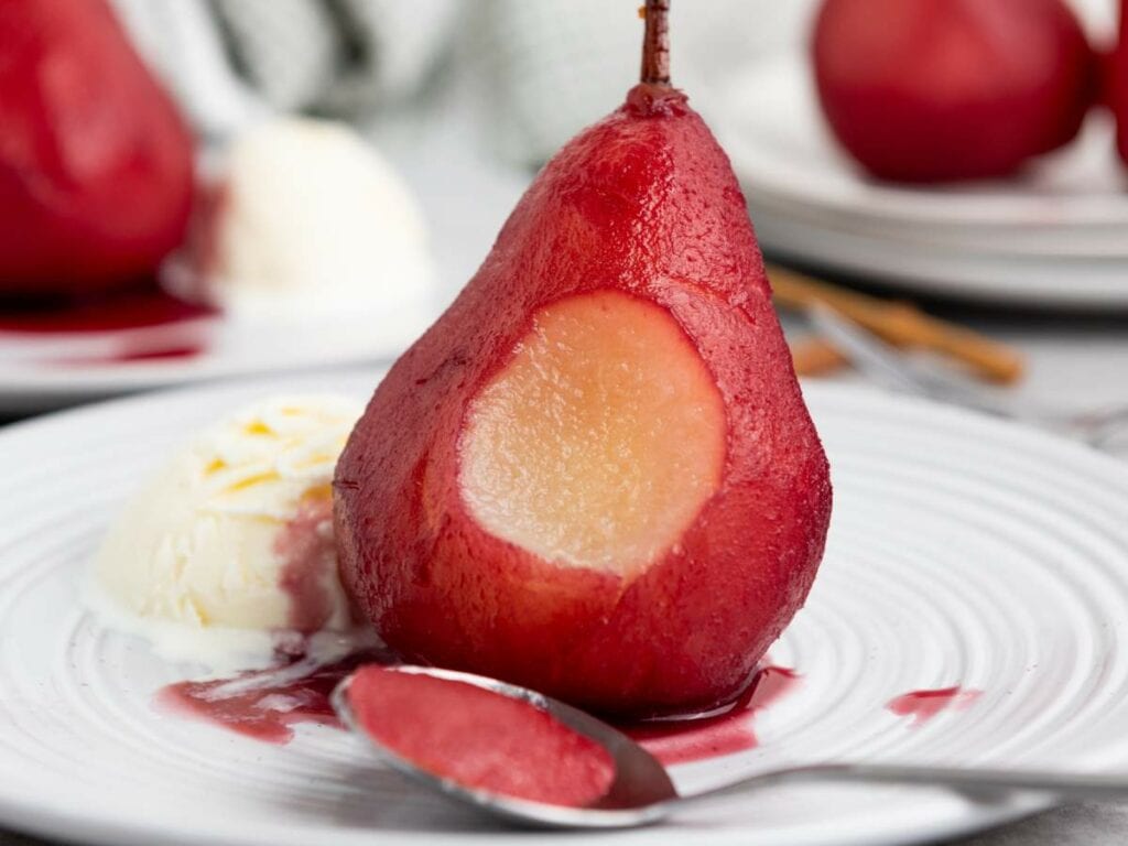 A poached pear with a bite taken out, served with a scoop of vanilla ice cream and a spoon on a white plate.