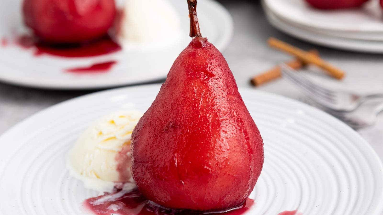 A poached pear in red sauce served with a scoop of vanilla ice cream on a white plate, with cinnamon sticks and a fork in the background.