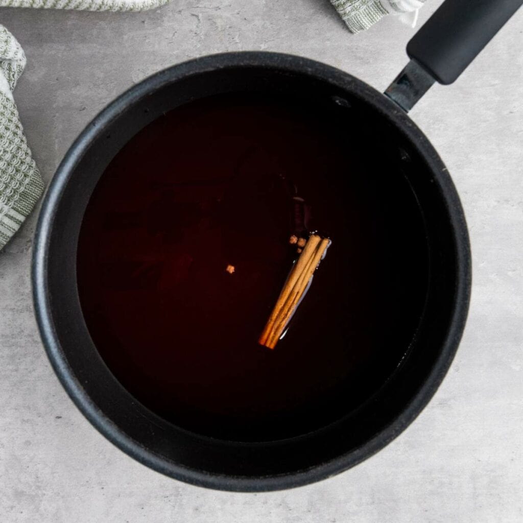 A saucepan filled with a dark liquid, containing a stick of cinnamon, sits on a light gray countertop near a textured towel.