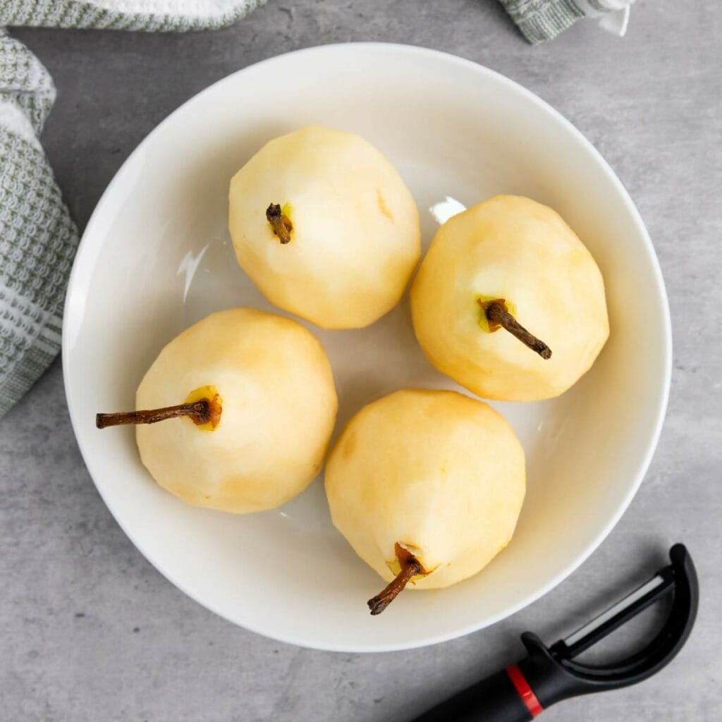 A white bowl containing four peeled pears with stems, placed on a gray surface next to a vegetable peeler and a cloth.