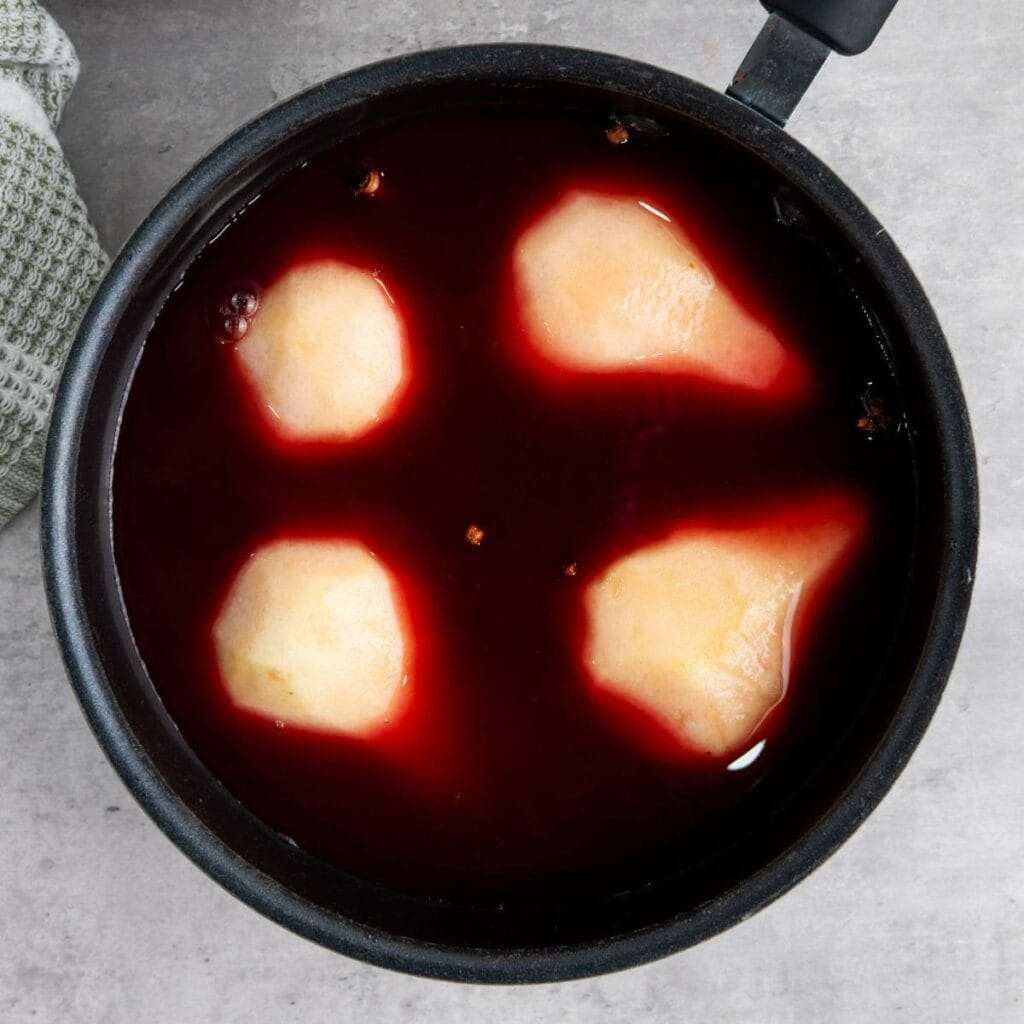 Four peeled pears are poaching in a pot of dark red liquid, likely wine or juice, viewed from above on a gray surface.