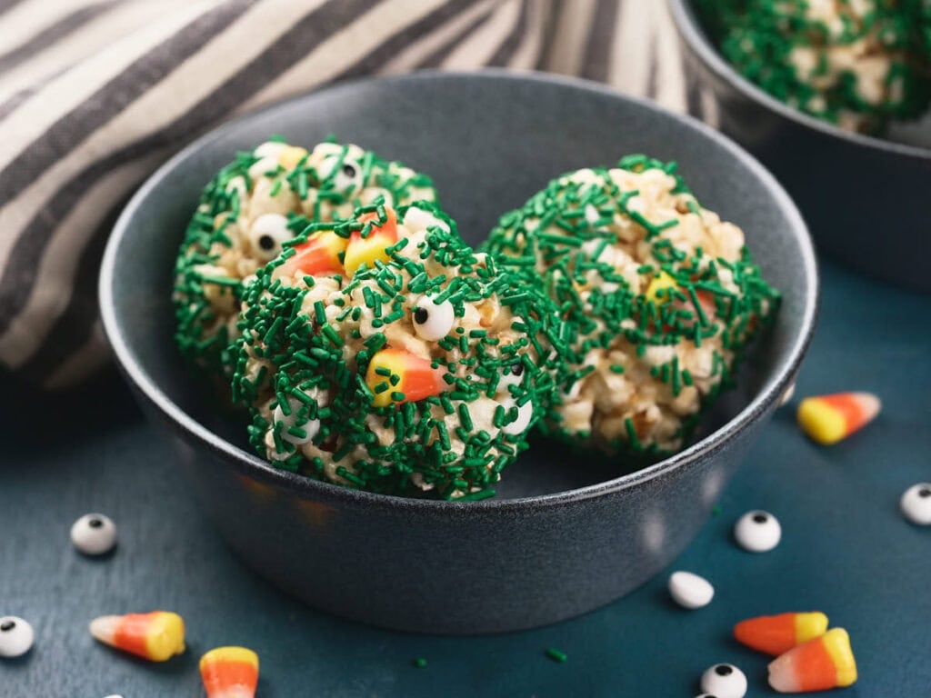 A bowl contains three popcorn balls covered in green sprinkles, candy eyes, and candy corn on a blue surface.