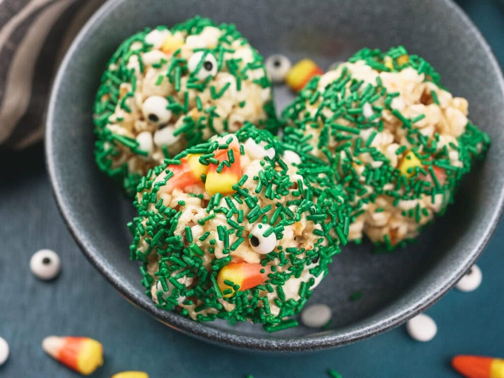 Three round popcorn treats covered in green sprinkles, candy eyes, and candy corn, served in a grey bowl.