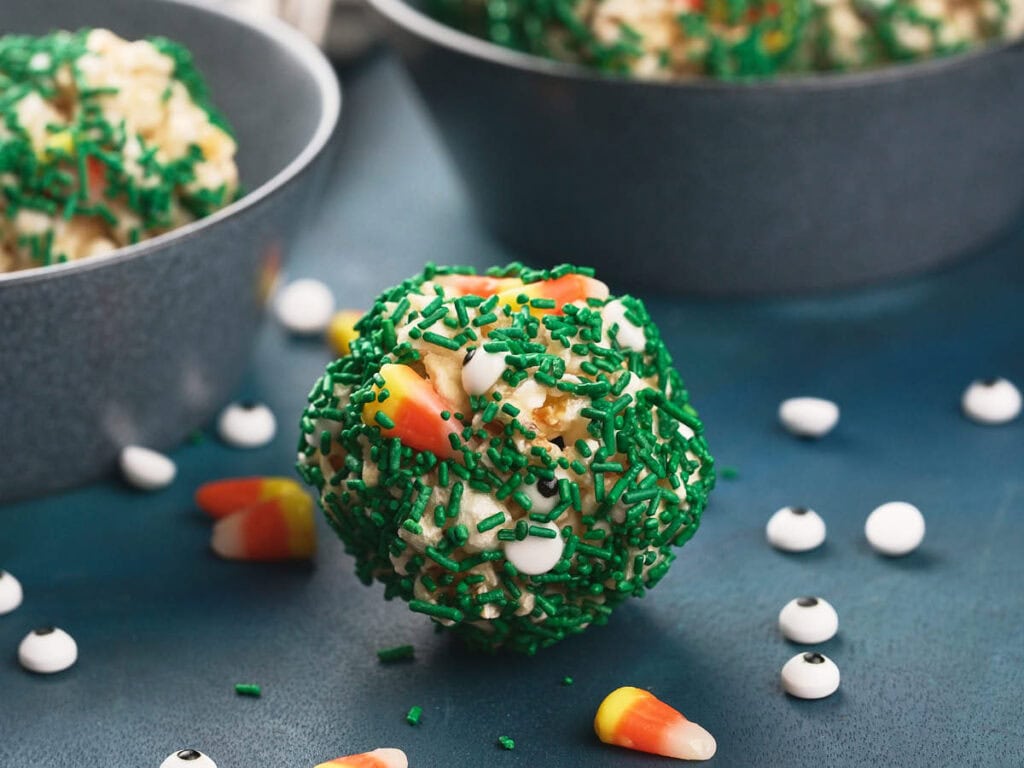 A round popcorn ball covered in green sprinkles, candy corn, and small candy eyeballs sits on a blue surface.