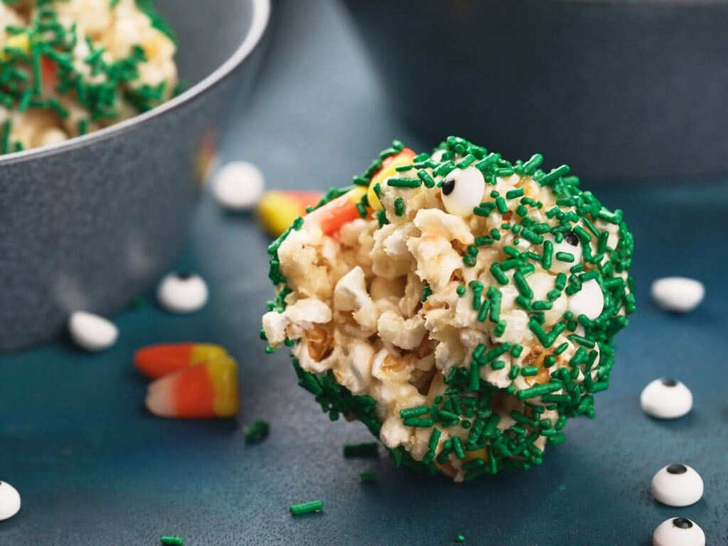 A popcorn ball covered in green sprinkles sits on a blue surface, with candy corn and candy eyeballs scattered nearby.