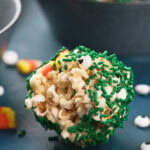 A popcorn ball coated with green sprinkles and small candy eyes, with candy corn pieces scattered nearby on a blue surface.