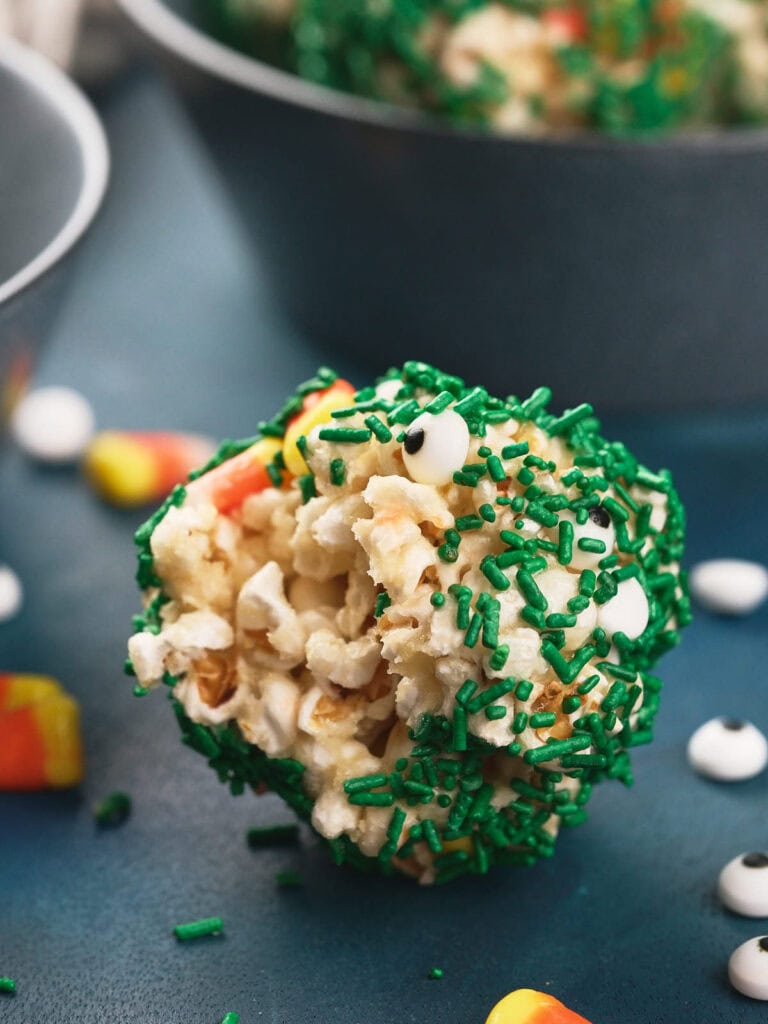 A popcorn ball coated with green sprinkles and small candy eyes, with candy corn pieces scattered nearby on a blue surface.