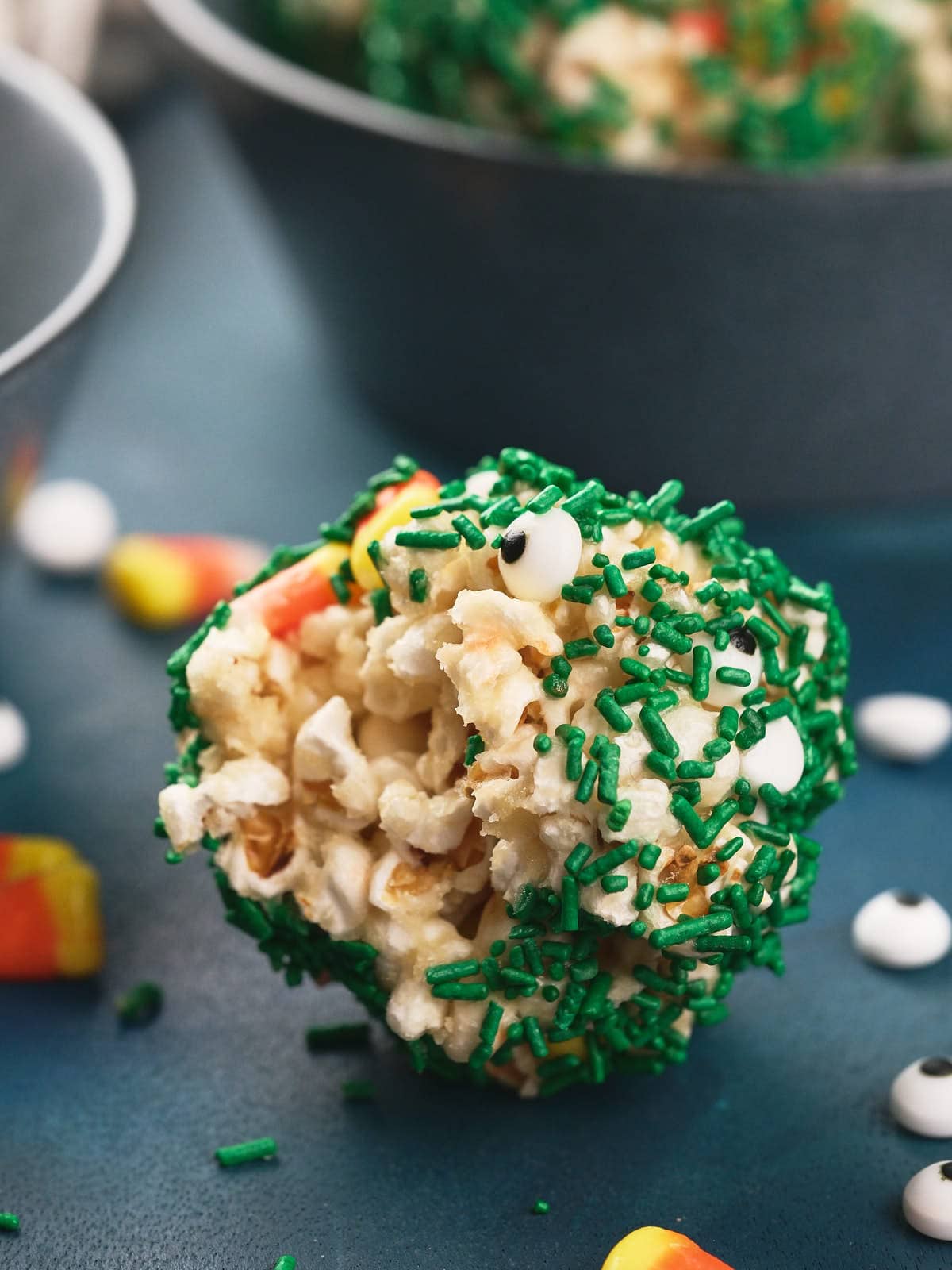 A popcorn ball coated with green sprinkles and small candy eyes, with candy corn pieces scattered nearby on a blue surface.