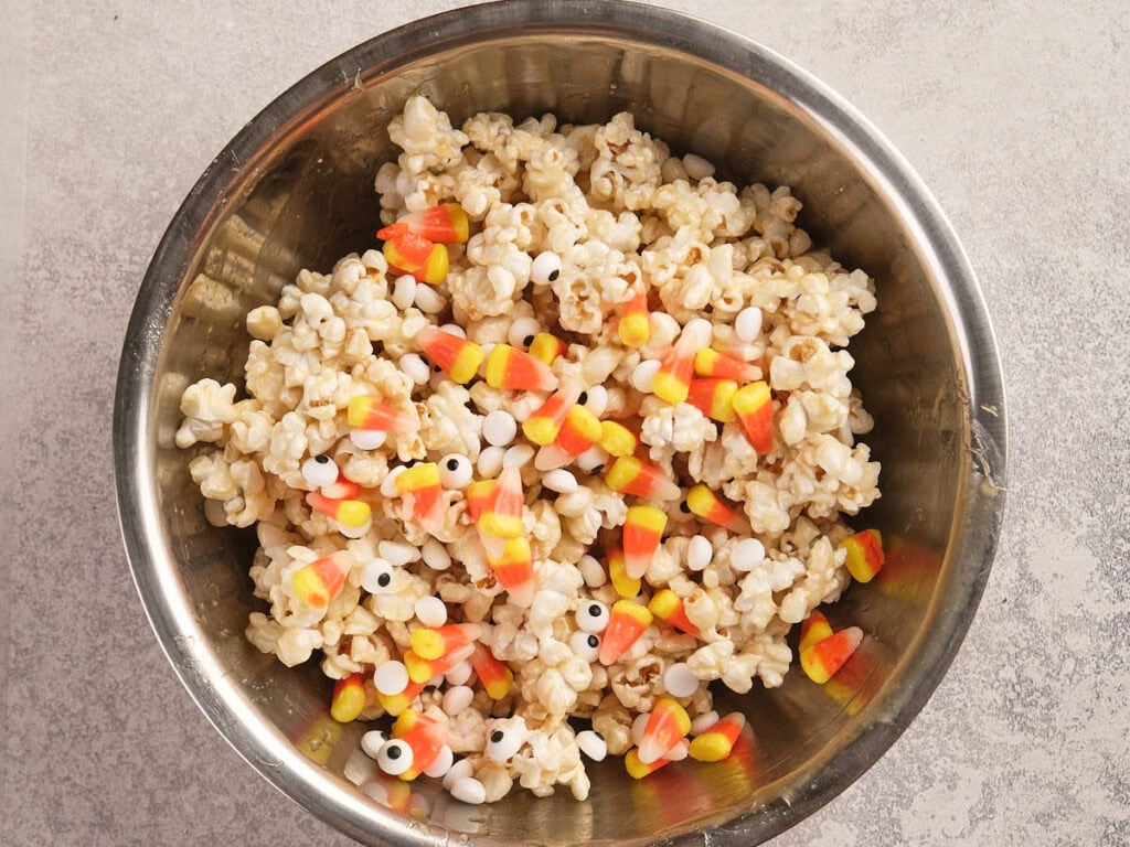 A metal bowl filled with popcorn, candy corn, and small white candy eyeballs, placed on a light-colored surface.