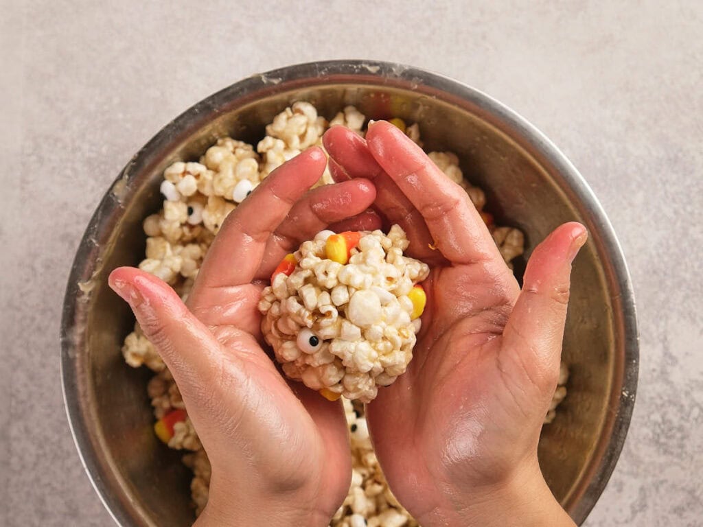 Hands shaping a popcorn ball mixed with candy corn and candy eyes over a metal mixing bowl.