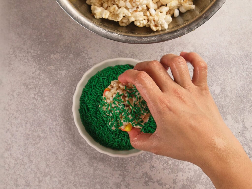 A hand dips a popcorn ball into a bowl of green sprinkles.