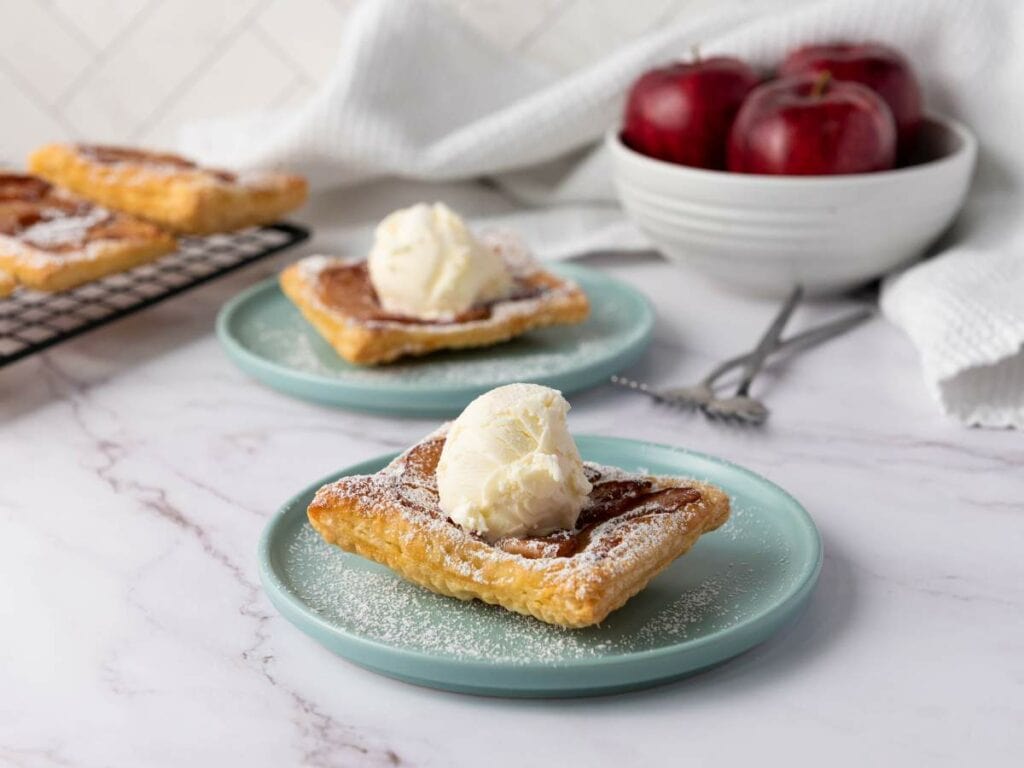 Two square apple tart topped with a scoop of vanilla ice cream on light blue plates.