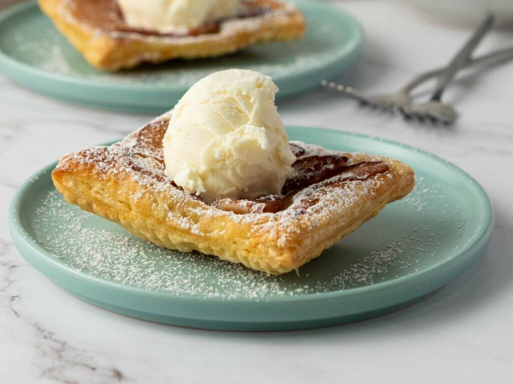 An apple tart topped with a scoop of vanilla ice cream and dusted with powdered sugar sits on a light blue plate.