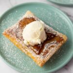 A square puff pastry topped with a scoop of vanilla ice cream and dusted with powdered sugar on a light blue plate.