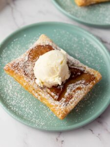 A square puff pastry topped with a scoop of vanilla ice cream and dusted with powdered sugar on a light blue plate.