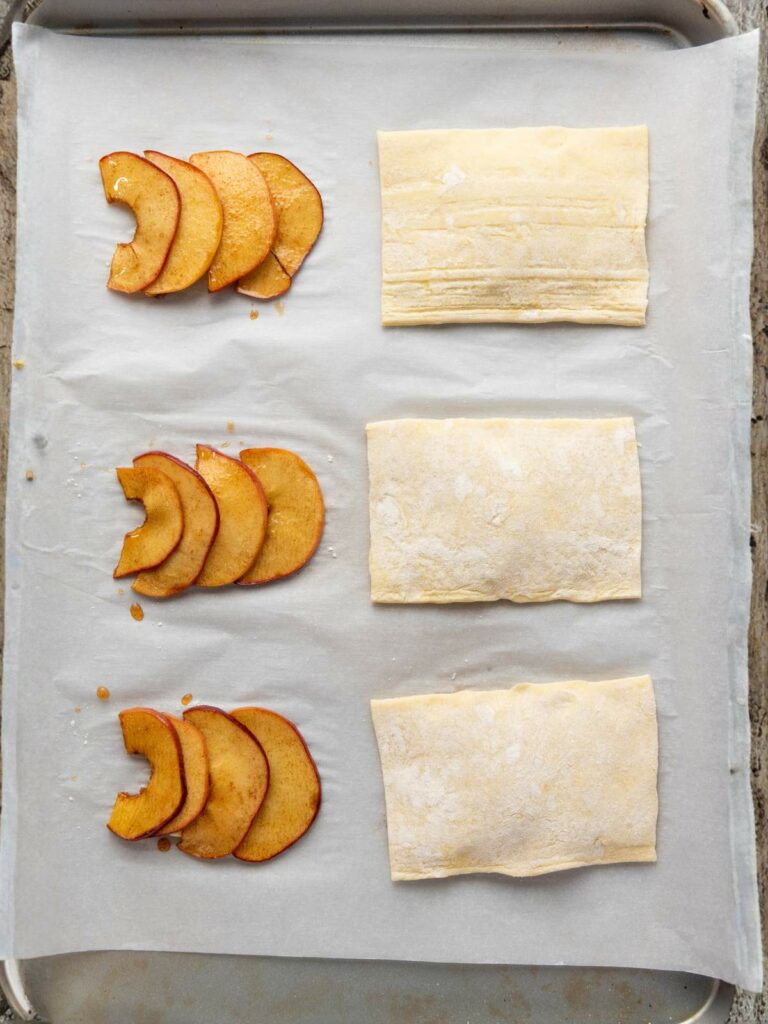 A baking sheet with three rows of thinly sliced apples arranged beside three rectangular pieces of puff pastry on parchment paper.