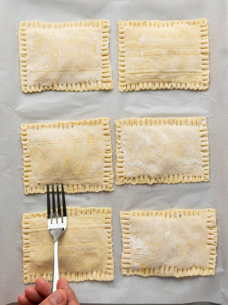 Six uncooked rectangular pastries on parchment paper; a hand presses a fork along the edge of one to seal it.