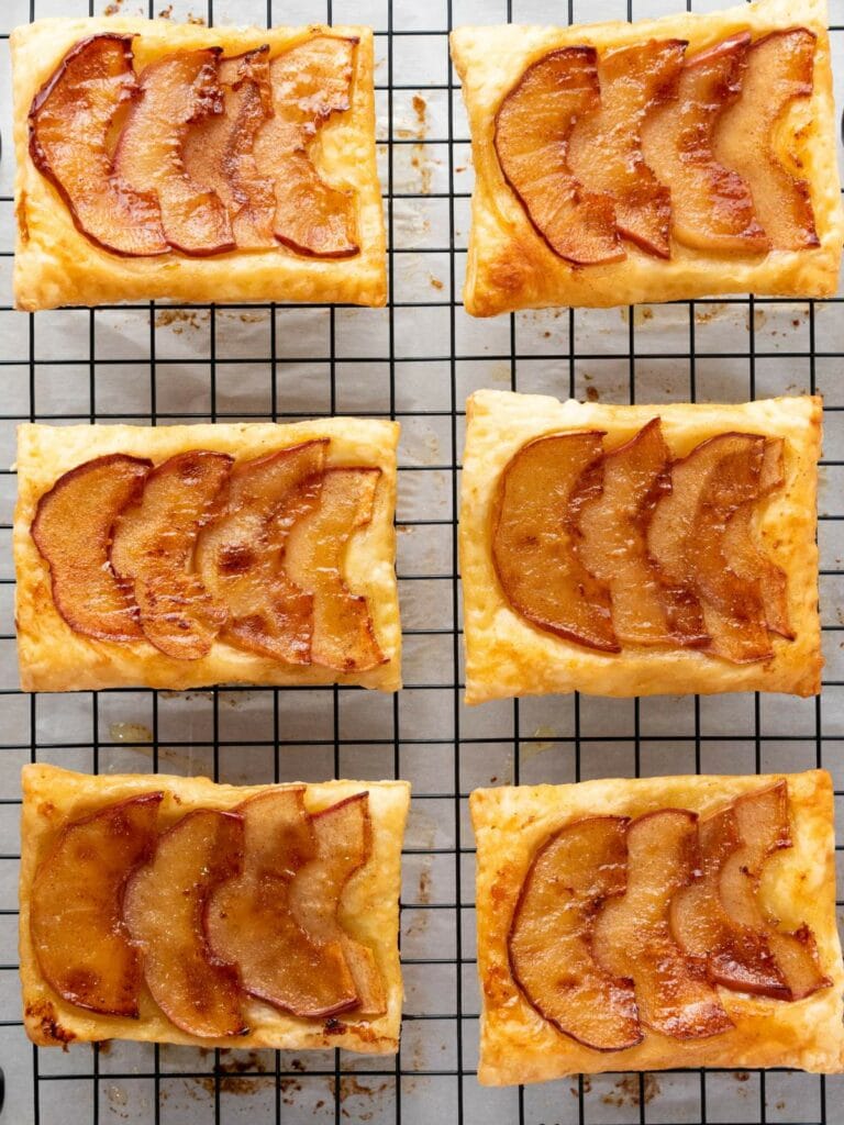 Six rectangular apple tarts with caramelized apple slices on top are cooling on a black wire rack.