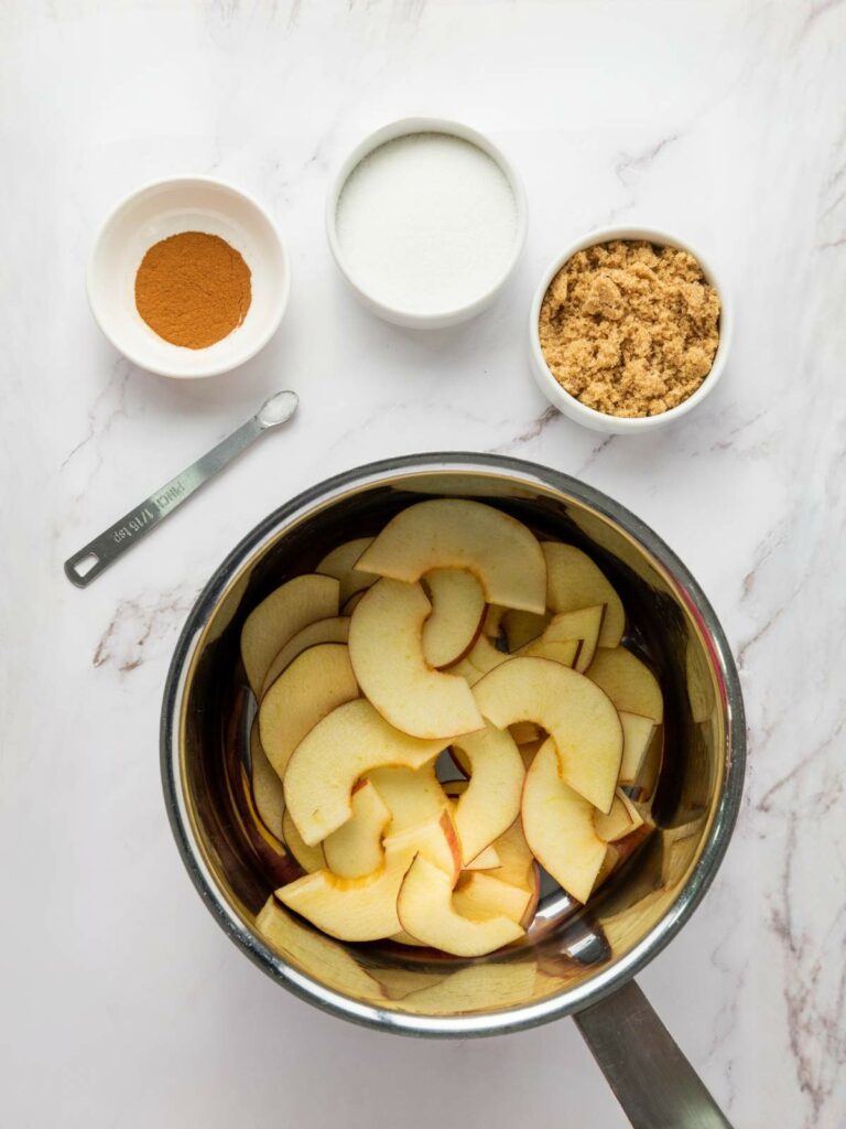 A saucepan with sliced apples, next to small bowls of cinnamon, white sugar, brown sugar, and a measuring spoon on a marble surface.