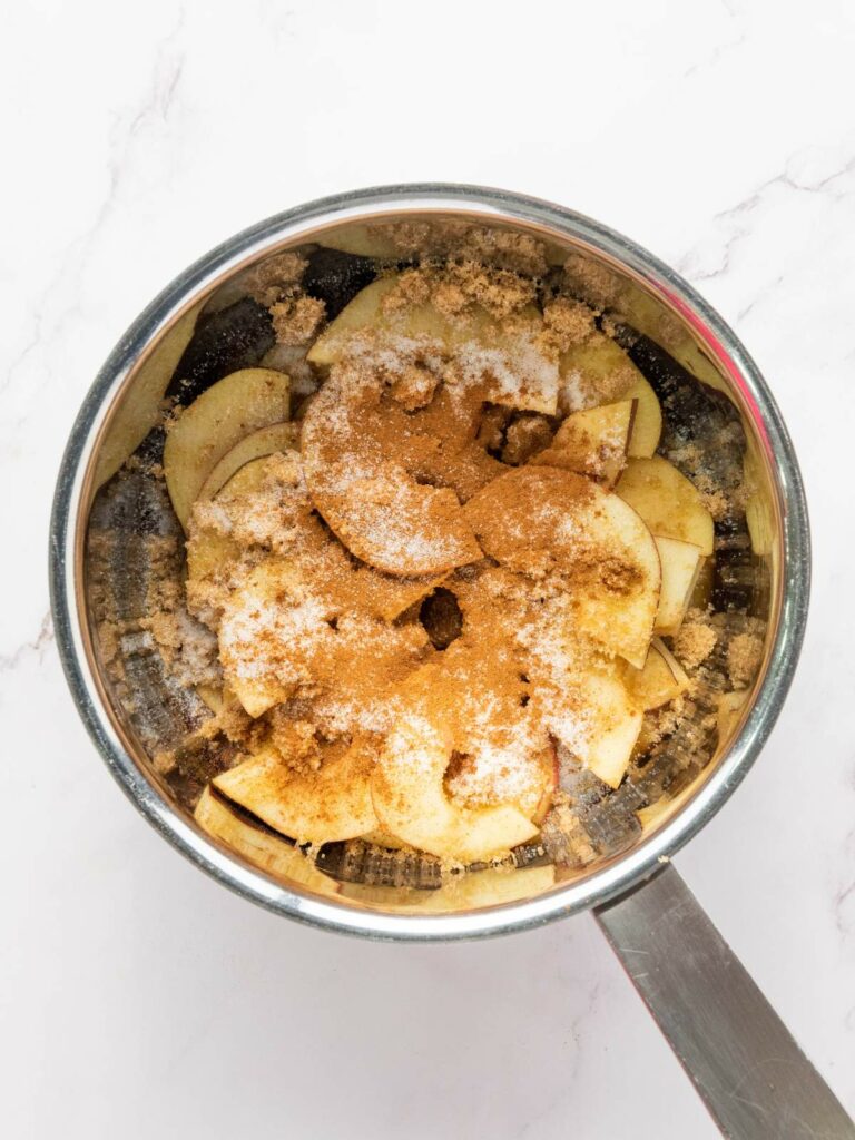 A saucepan containing sliced apples topped with brown sugar, white sugar, and cinnamon, resting on a white marble surface.